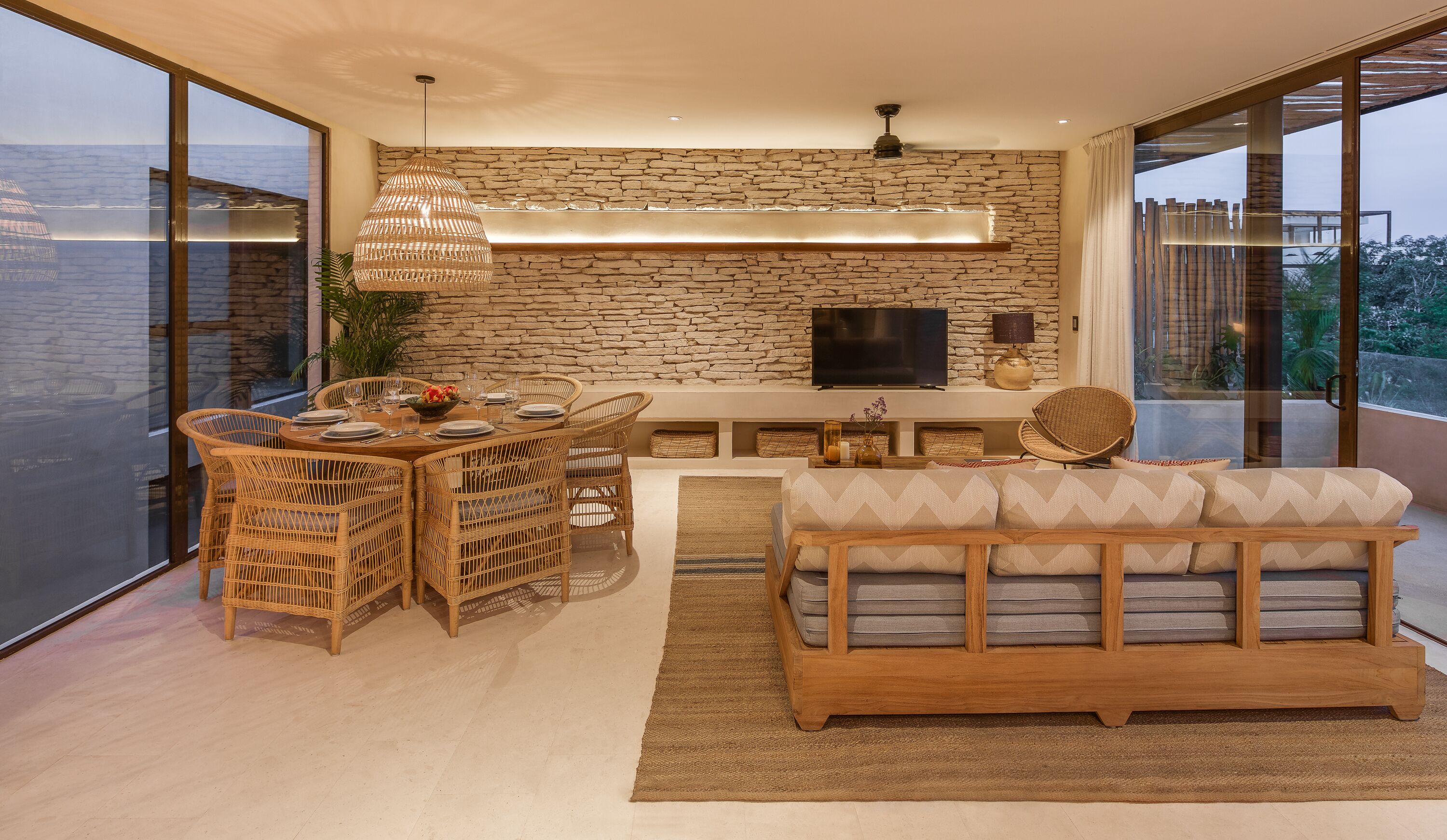 A modern living room with a stone wall, a flat-screen TV, wicker furniture, and a large window.