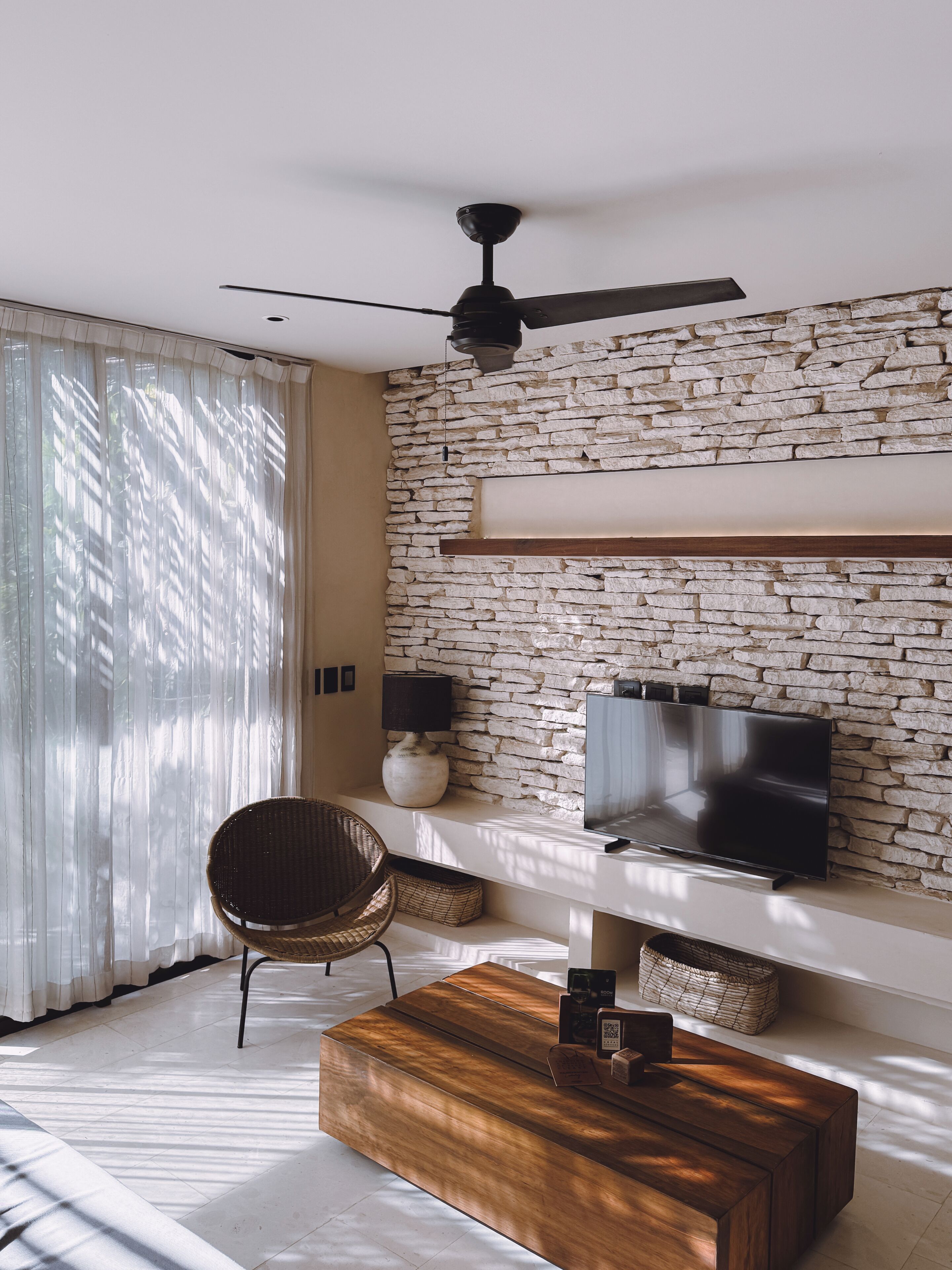 Una sala de estar moderna con pared de piedra, un televisor de pantalla plana, un ventilador de techo y una mesa de centro de madera.