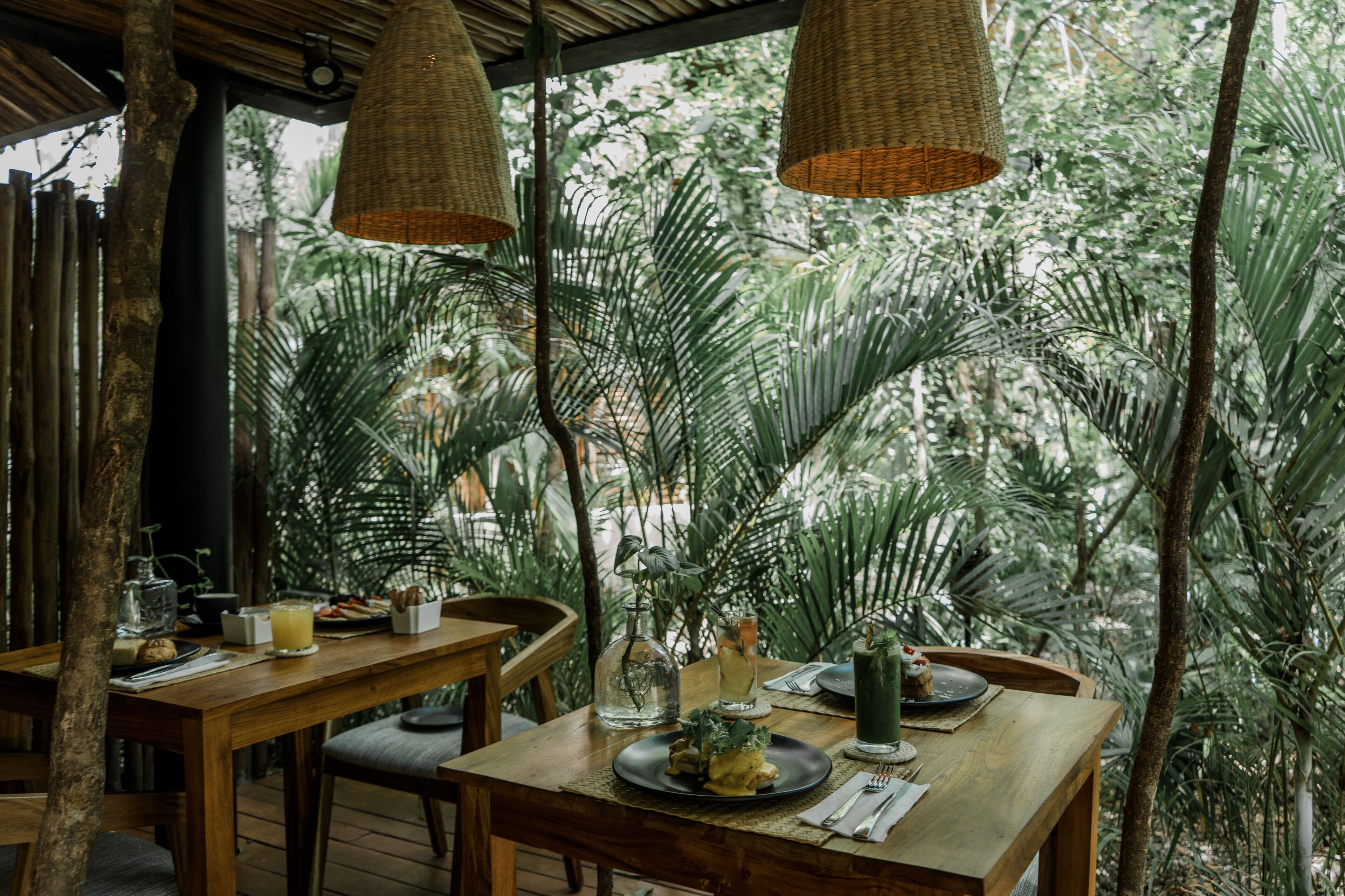 Una mesa de comedor de madera dispuesta con platos, vasos y una botella, rodeada de sillas en un entorno tropical con lámparas de mimbre colgantes.