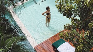 A woman in a black bikini standing in a clear pool surrounded by lush greenery.