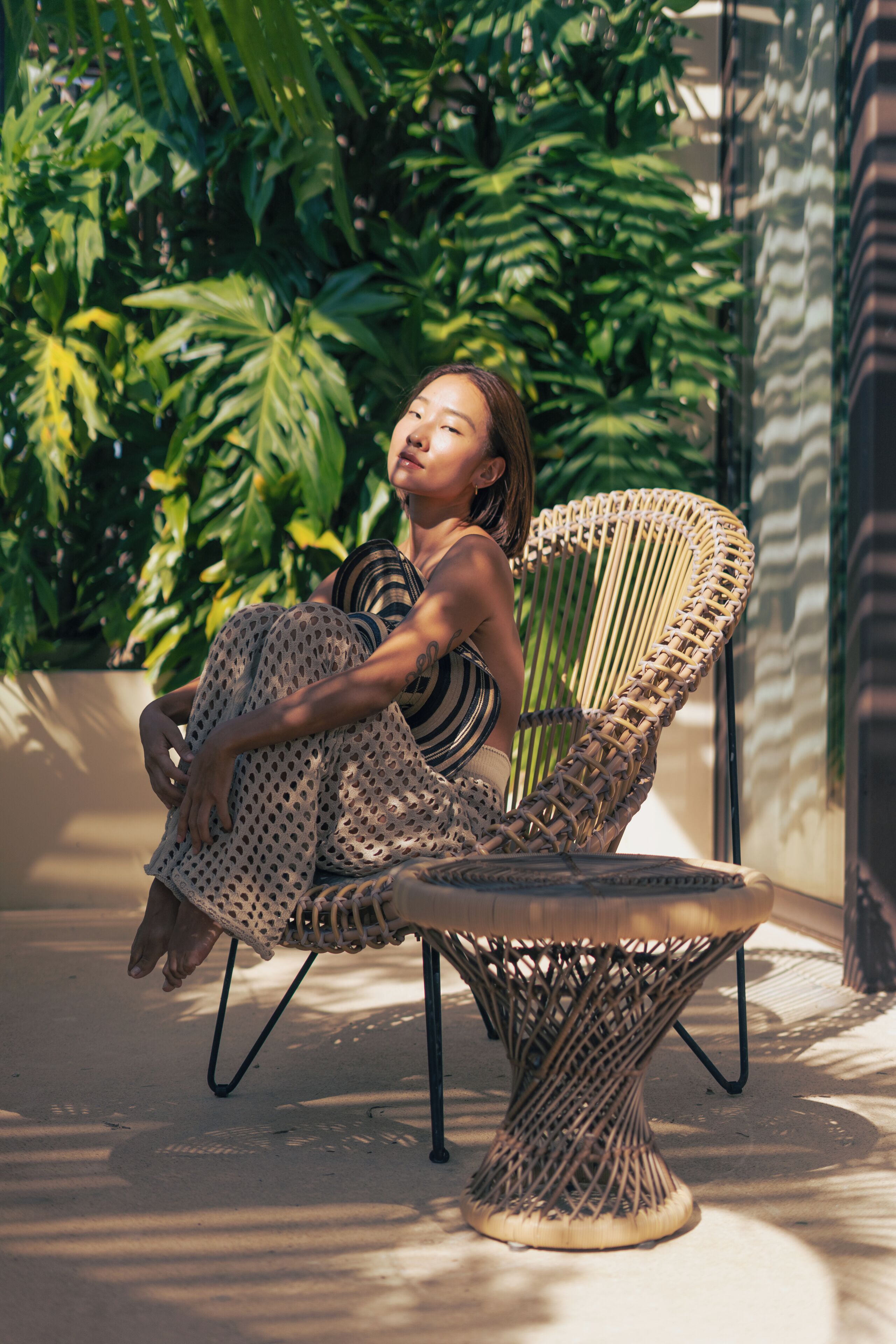 A person sitting on a wicker chair with a woven base, outdoors with greenery in the background.