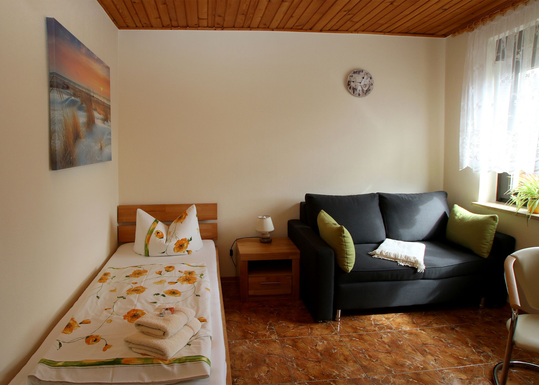 Un dormitorio con cama, mesita de noche, sofá, sillón, reloj y ventana con cortinas.