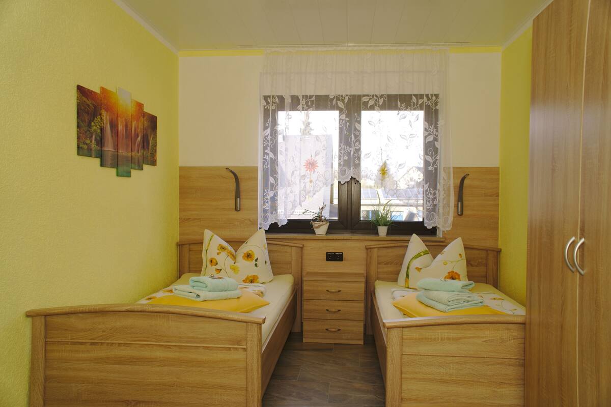 A room with two wooden beds, a window with floral curtains, and a small desk with a potted plant.