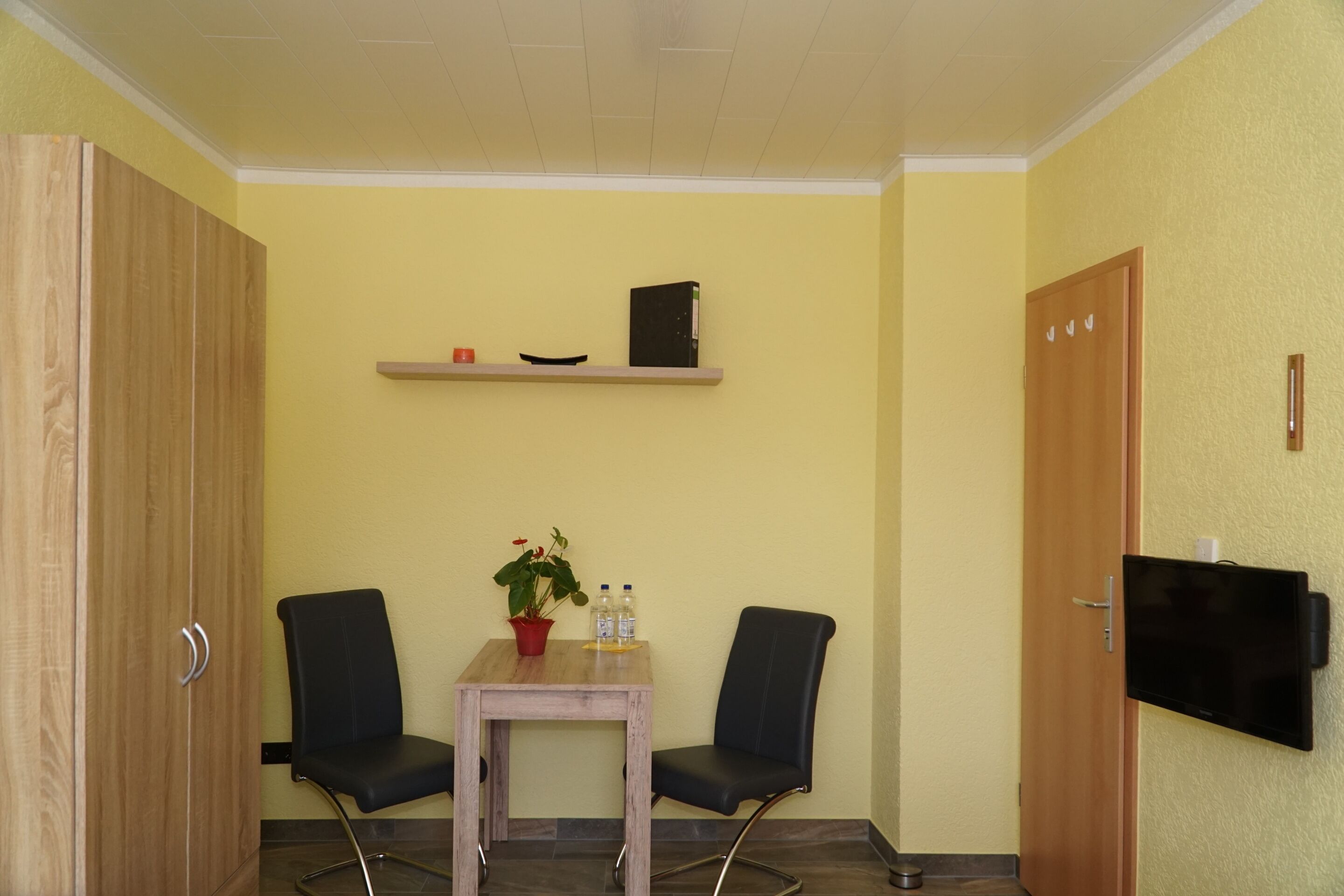 A small room with a wooden wardrobe, a table with chairs, a shelf with a book and a bowl, a potted plant, and a TV mounted on the wall.