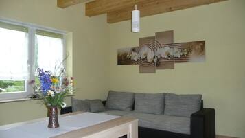 Ein Wohnzimmer mit grauem Sofa, einem Holztisch und einer Vase mit Blumen. Es gibt ein Fenster mit Vorhängen und ein an der Wand befestigtes Kunstwerk über dem Sofa.