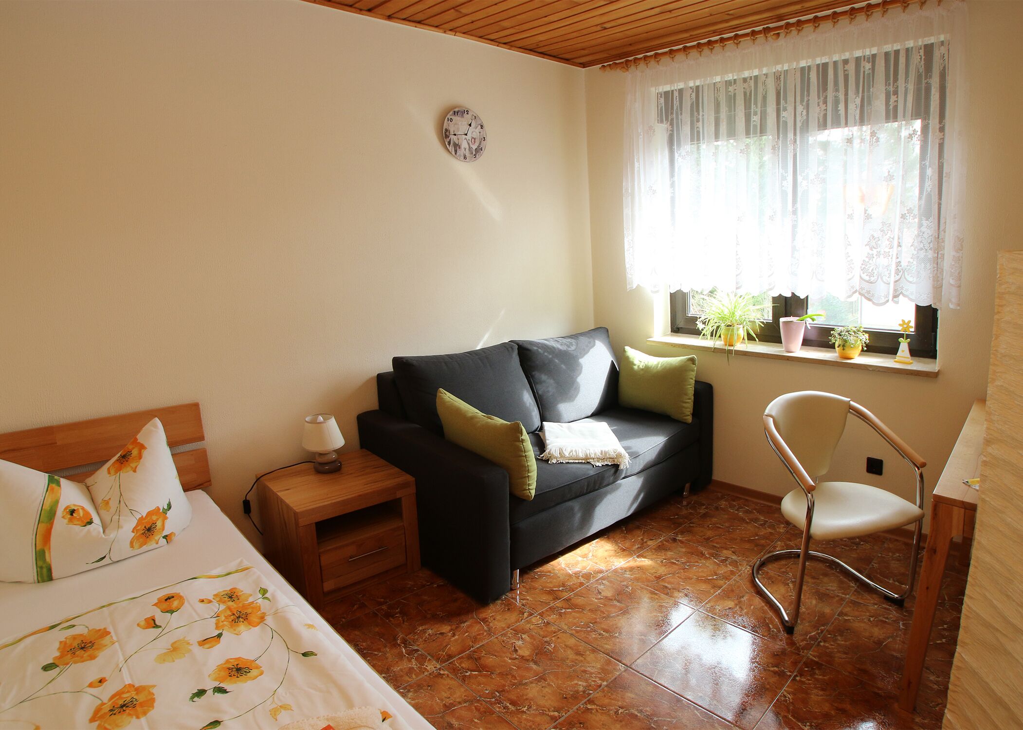 A cozy room with a bed, a dark-colored sofa with cushions, a small wooden nightstand with a lamp, a chair, and a window with curtains.