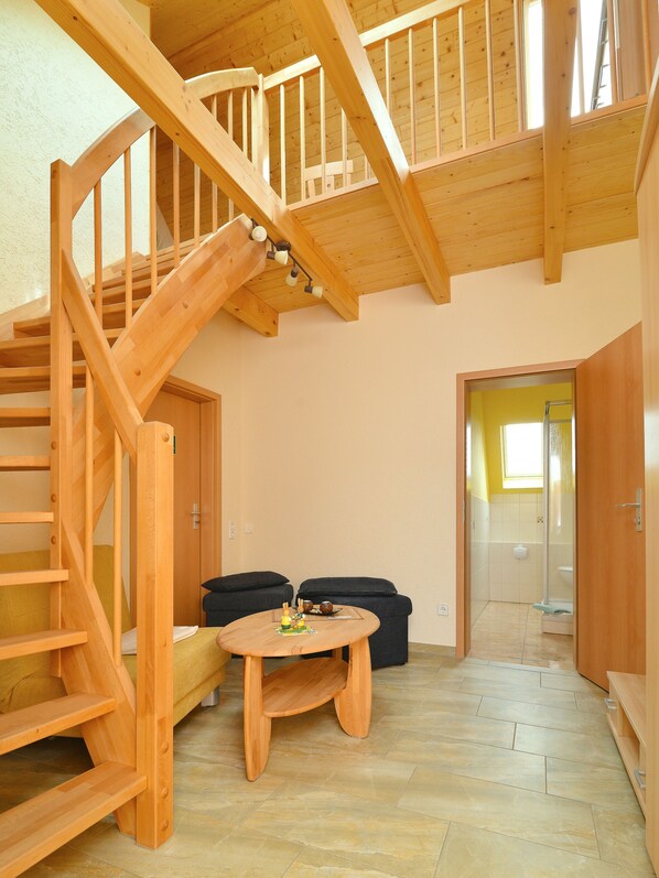 A wooden staircase leading to an upper level, a small round table with a cushion, and a doorway to a bathroom. - Pension Andrea (Lugau)