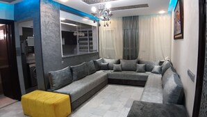 Ein modernes Wohnzimmer mit einem grauen Ecksofa, einem gelben Pouf und einer Kronleuchter.