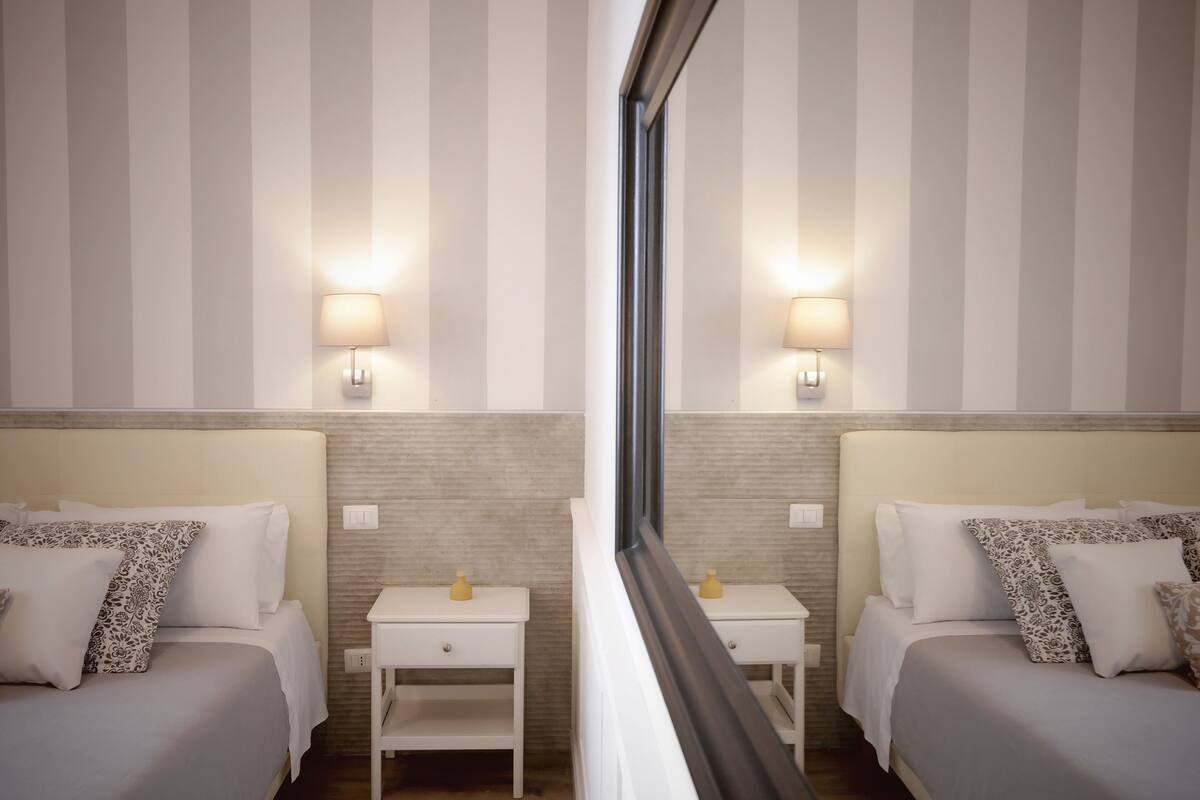Ein Hotelzimmer mit zwei Betten, jeweils mit weißem Kopfteil und passenden Kissen. An den Betten stehen Nachttische mit Lampen und Spiegeln. Die Wände sind mit vertikalen Streifen versehen, und ein Fenster bietet Blick ins Zimmerinnere.