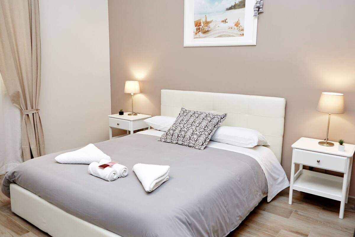 Ein ordentlich bezogenes Bett mit grauer Tagesdecke, weißen Kissen und gerollten Handtüchern. Zwei Nachttische mit Lampen, eine kleine Pflanze und ein gerahmtes Bild an der Wand.