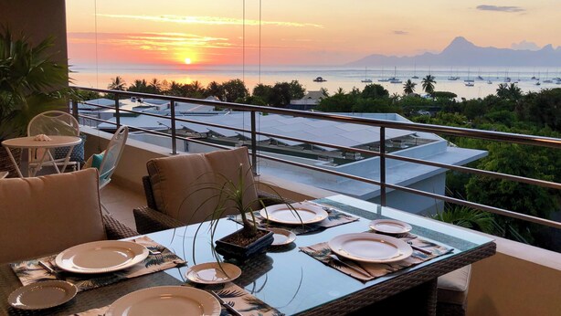 Un coin repas avec une table dressée pour quatre personnes, offrant une vue sur un magnifique coucher de soleil, avec des bateaux et des montagnes en toile de fond.