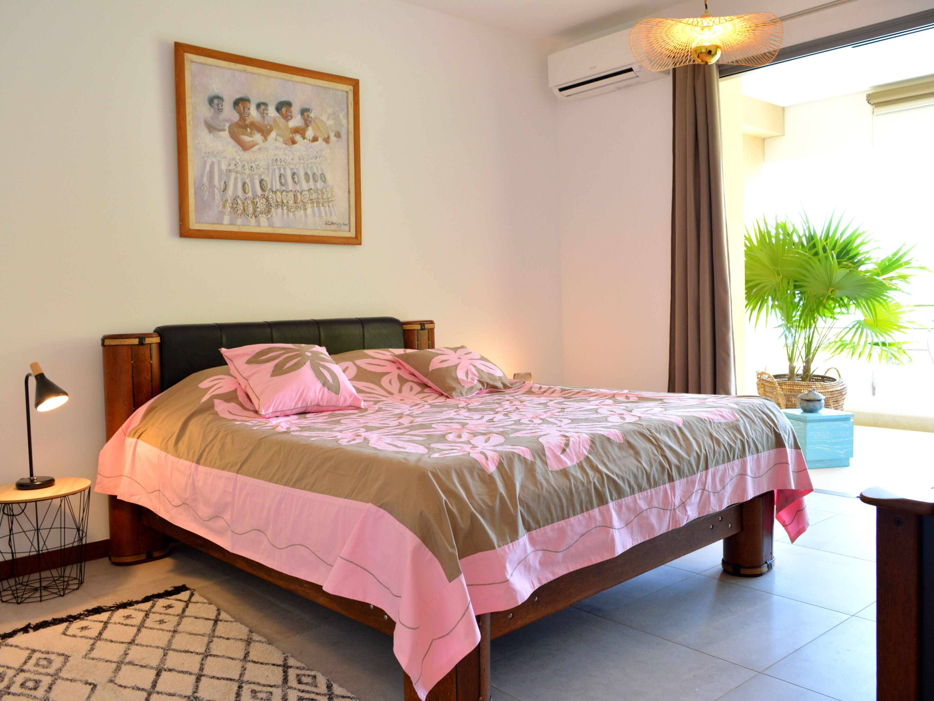 A bedroom with a bed, a bedside table, a lamp, a framed picture on the wall, an air conditioning unit, and a potted plant.