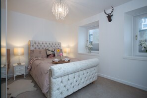 Une chambre à coucher avec une tête de lit capitonnée, un lustre, une décoration représentant une tête de cerf, une table de chevet avec une lampe et une fenêtre offrant une vue.