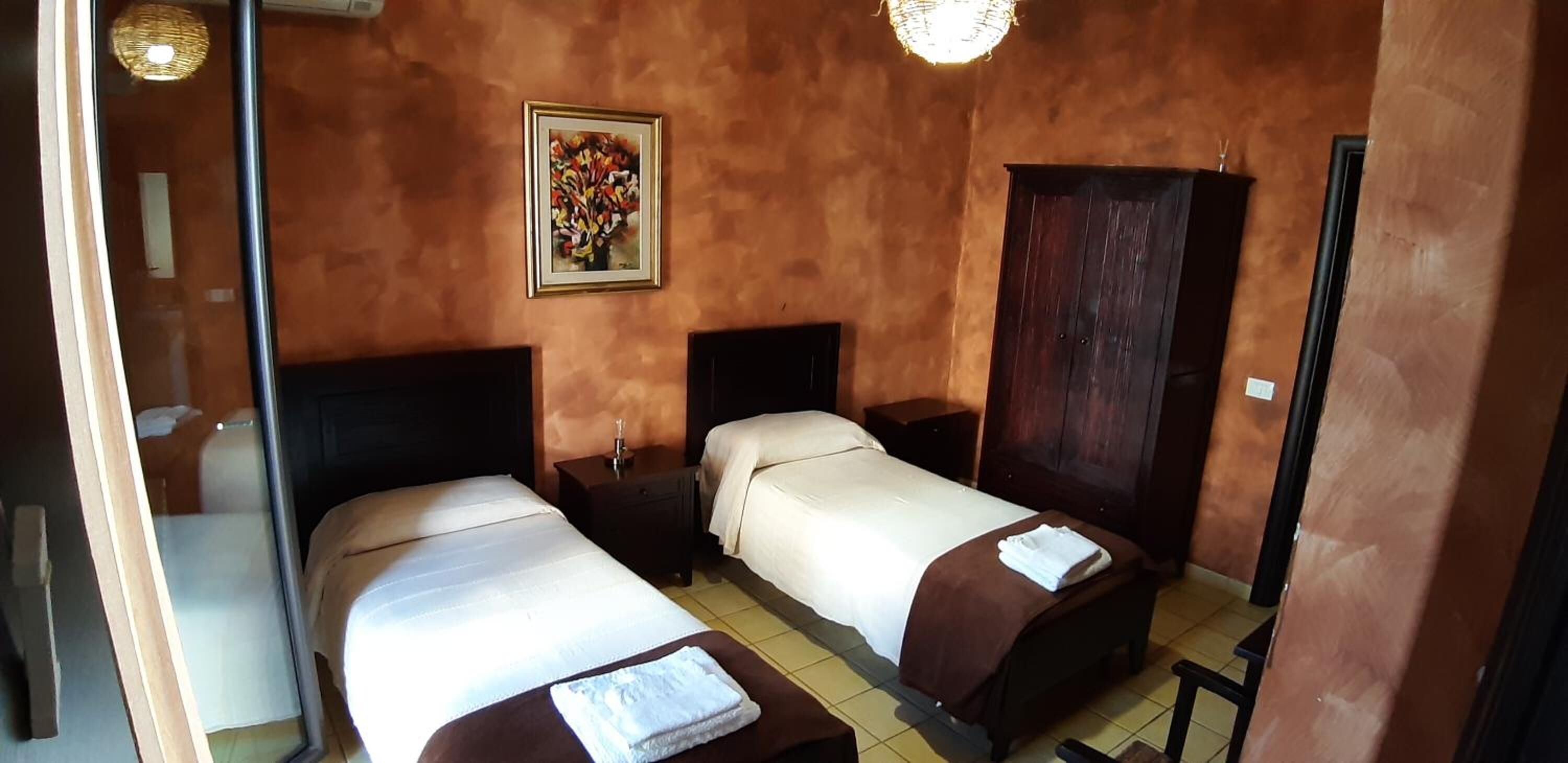A room with two beds, a wooden wardrobe, a painting on the wall, and a small table with a lamp.