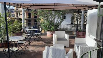 Eine Terrassengruppe im Freien mit runden Tischen und weißen Korbstühlen, umgeben von Topfpflanzen und einer Pergola.