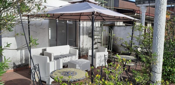 Un patio con mobiliario exterior, que incluye un sofá y sillas, bajo un amplio paraguas.