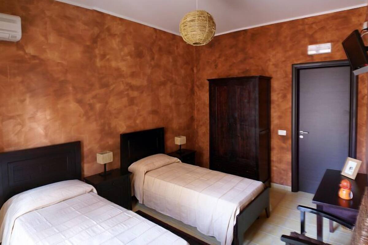 Una habitación con dos camas, un armario, un escritorio con silla, una lámpara colgante y una puerta.