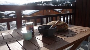 Ett träbord med ett bröd, en skål och en burk på en balkong med utsikt över ett snötäckt landskap med hus och berg.