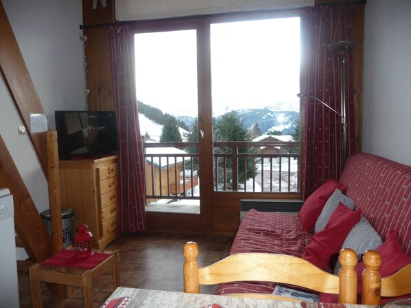 A room with a wooden bed, a television on a wooden cabinet, a small table with a red lantern, and a view of snowy mountains through a balcony door. - Savoie, Col des Saisies, Apartment for rent (Hauteluce)