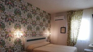 A bedroom with floral wallpaper, a bed with a beige bedspread, a nightstand, a wall-mounted air conditioner, and a window with curtains. - Aramoni B&B (Zambrone)