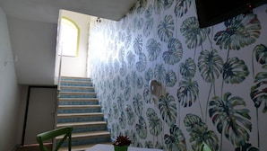 A room with a staircase, a green chair, a table with a potted plant, and a wall with a tropical leaf pattern. - Aramoni B&B (Zambrone)