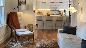 Ein Wohnzimmer mit einer Couch, einem Hocker und einem Sessel. Im Hintergrund ist eine Küchenzeile mit einer Bar und einer Pendelleuchte zu sehen.