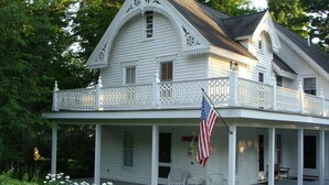 A white colonial-style house with a balcony, an American flag, and a porch with wicker chairs. - Lake Chautauqua Victorian (Dewittville)