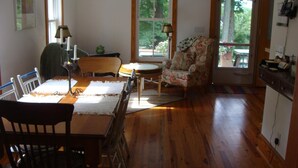 A dining area with a wooden table, chairs, and a floral armchair. There are windows with curtains, a lamp, and a small table with a teapot. - Lake Chautauqua Victorian (Dewittville)