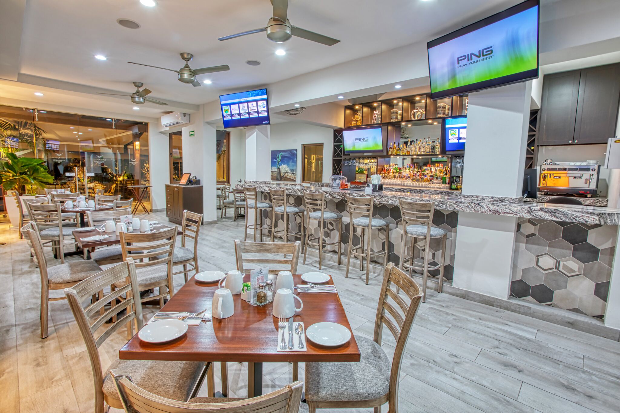 Un restaurant moderne doté de nombreuses tables et chaises, d’un bar avec un comptoir et des étagères remplies de bouteilles, et d’un grand écran affichant le nom de la marque, « PING ».