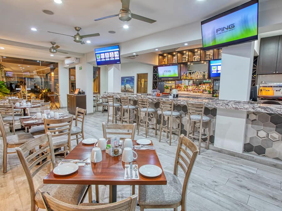 Un restaurante moderno con varias mesas y sillas, una zona de bar con mostrador y estantes llenos de botellas, y una pantalla grande que muestra el nombre de la marca 'PING'.