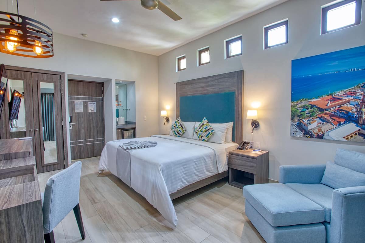 A modern hotel room with a large bed, a desk, a chair, a wardrobe, and a painting of a coastal city.