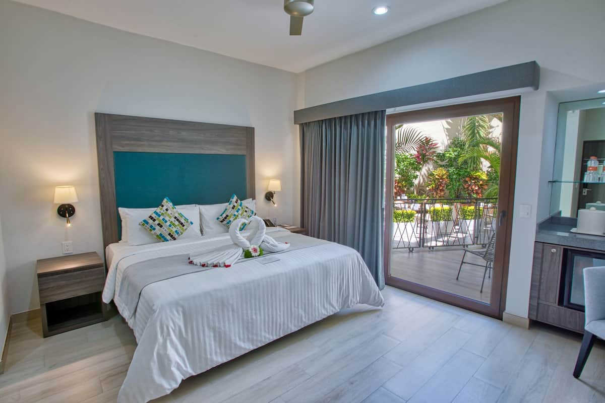A hotel room with a large bed, two bedside lamps, a view of a balcony with outdoor furniture, and a small kitchenette area.