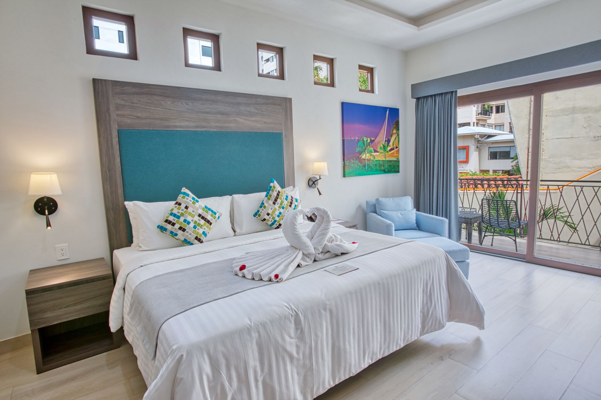 Une chambre d’hôtel avec un grand lit, une table de chevet, une chaise bleue et un balcon donnant sur des immeubles.