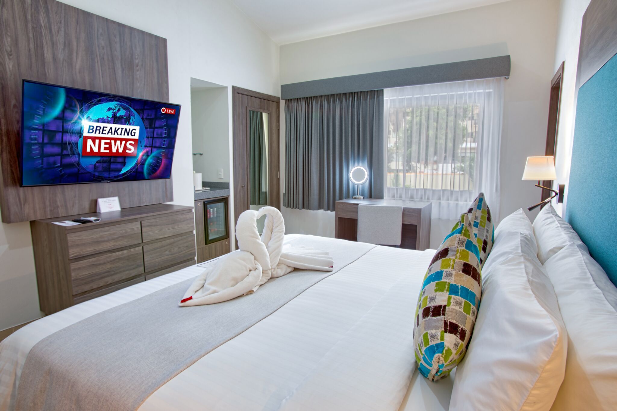 A hotel room with a large flat-screen TV displaying a news channel, a neatly made bed with white linens, a colorful pillow, a bedside table with a lamp, and a window with curtains.
