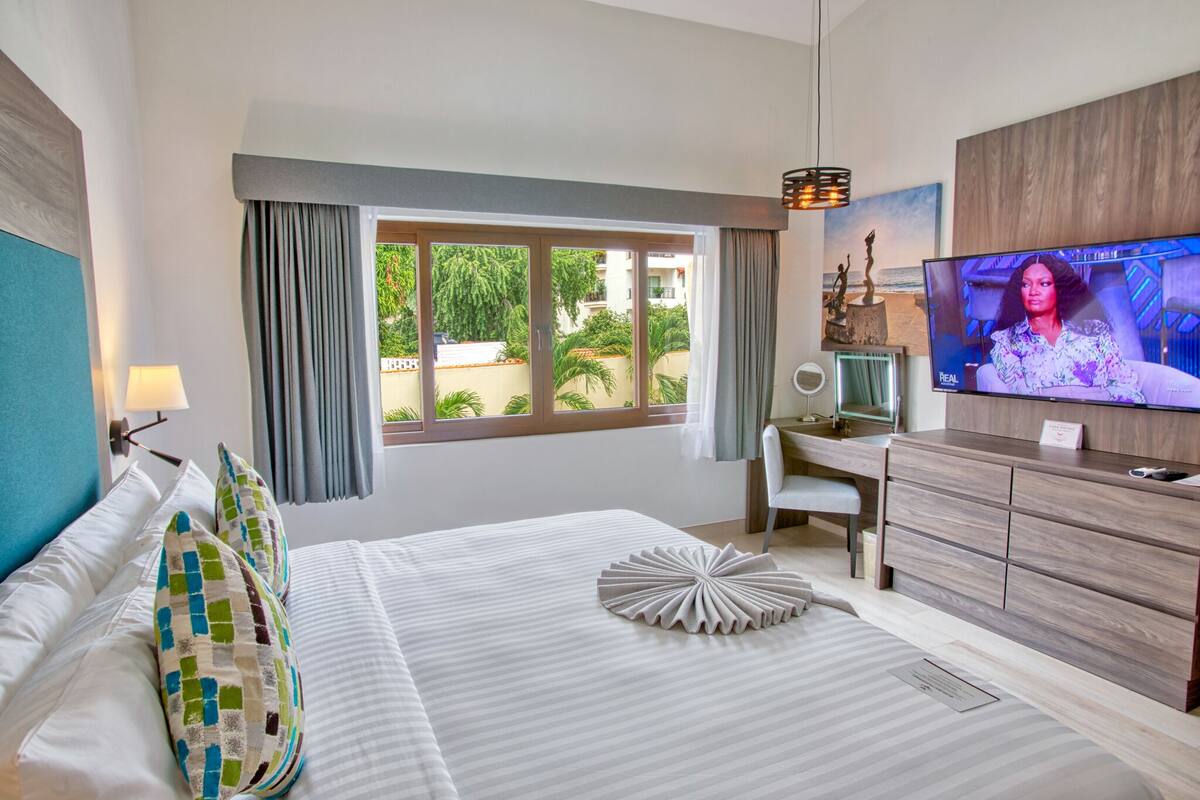 A hotel room with a bed, a desk, a TV, and a view of greenery outside.