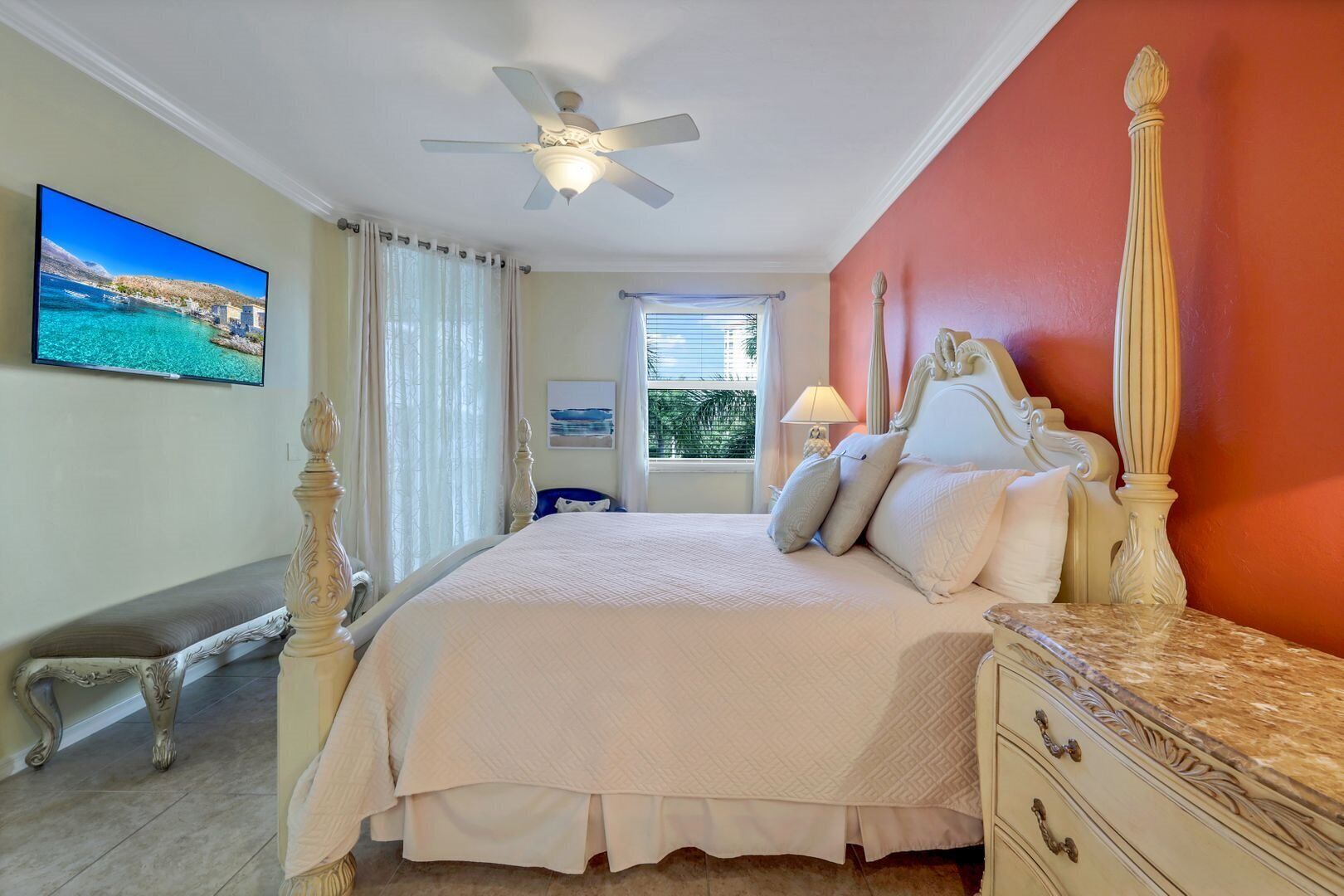 A bedroom with a large bed, a ceiling fan, a TV on the wall, and a window with curtains.