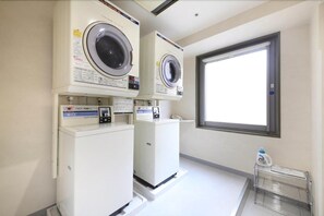 A laundry room with two front-loading washing machines, a small table with a bottle, and a window providing natural light. - R&B Morioka-ekimae (Morioka)