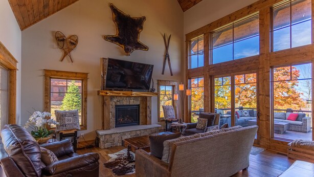 A spacious living room with a stone fireplace, leather sofas, and large windows offering a view of autumn foliage. - Red, White and Views at Eagles Nest- club amenities, hot tub, long range views, pet friendly (Banner Elk)