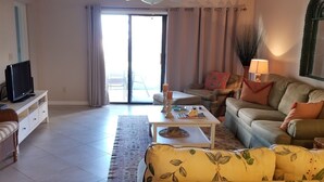 A living room with a sofa, a coffee table, a television on a stand, and a sliding glass door leading to an outdoor area. - 2 Bed, 2 Bath, Ocean View, Poolside - Sea Place 13137 (St. Augustine)