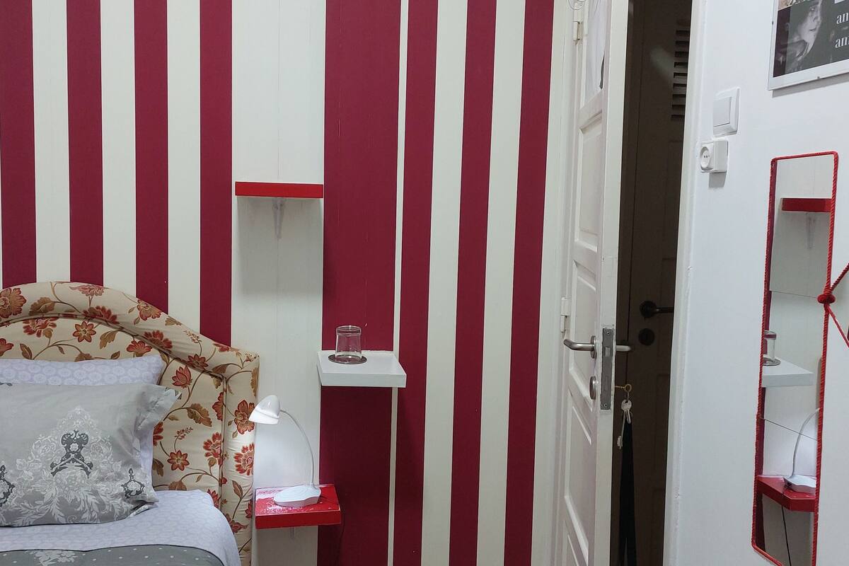A bedroom with a bed, a bedside table, a lamp, a mirror, and a door leading to another room.