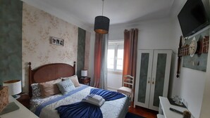 A bedroom with a bed, a bedside table, a lamp, a wardrobe, a TV, and a window with curtains. - Ameli Rooms (Lisbon)