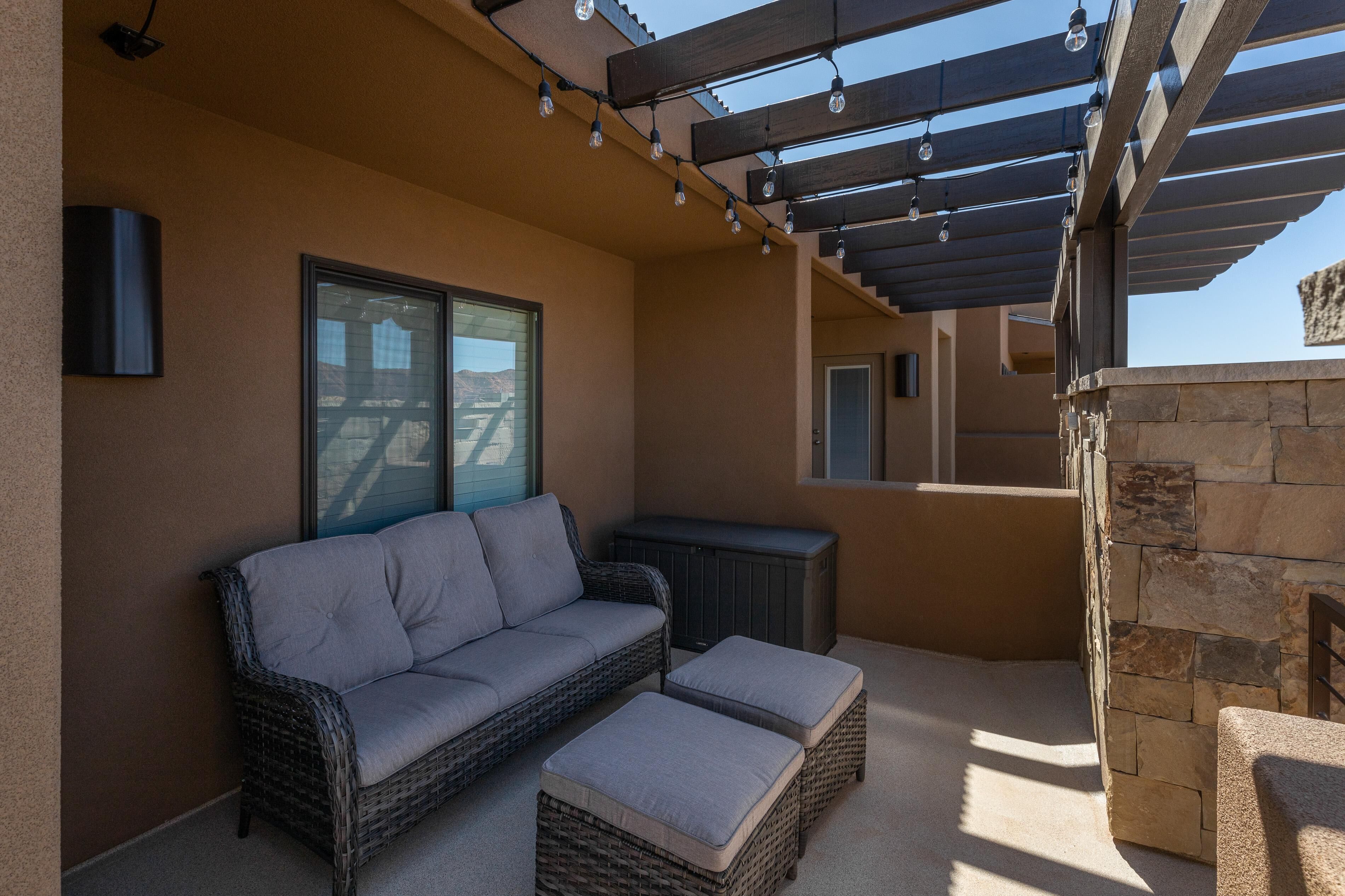 A covered patio area with a wicker sofa and ottoman, a stone fireplace, and a pergola with string lights.