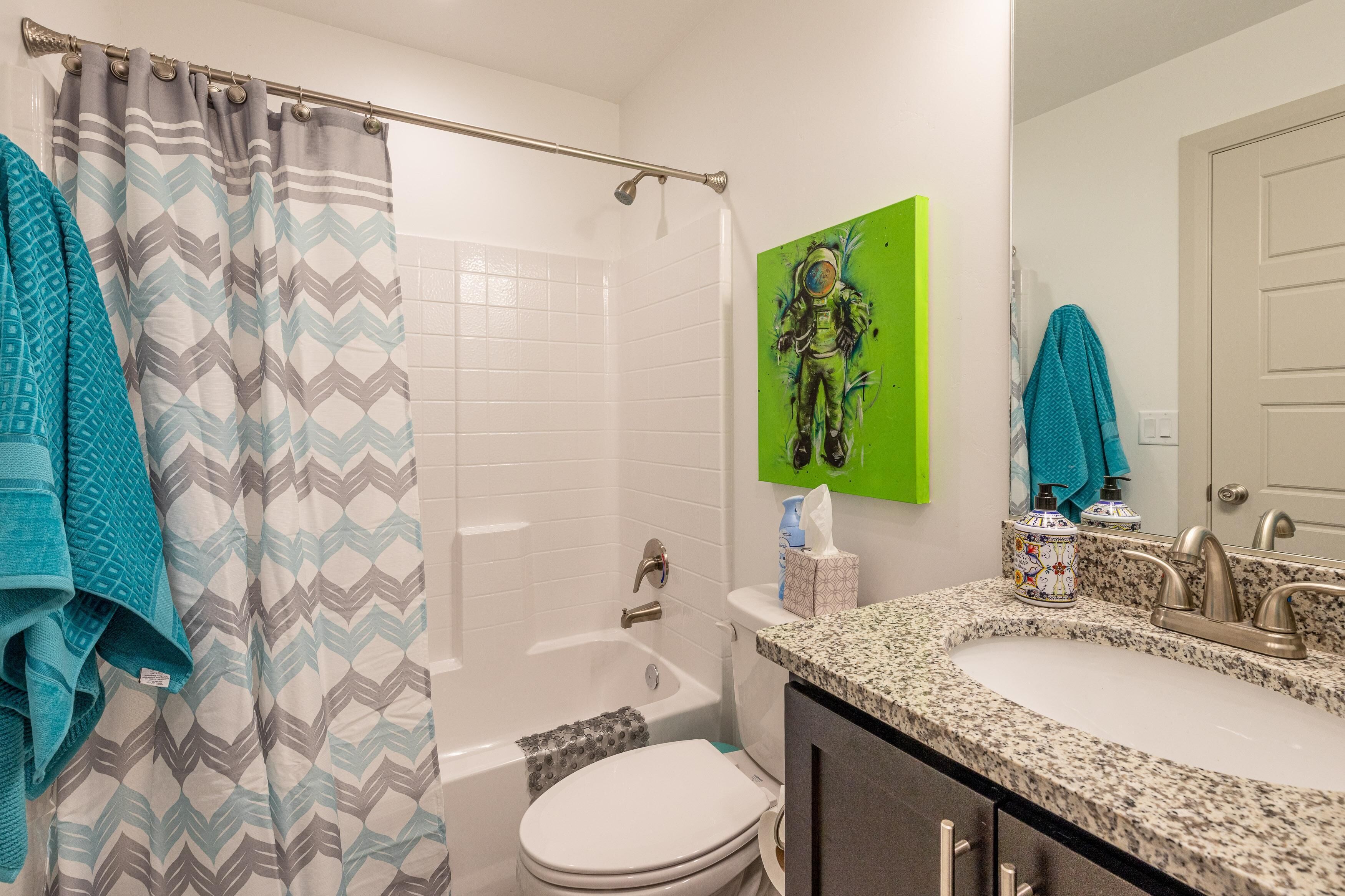 A bathroom with a shower curtain, a toilet, a sink, and a green wall art piece.
