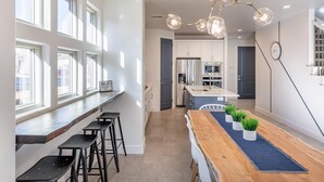 A modern kitchen with a long island, bar stools, and a dining area with a wooden table and potted plants. - Oasis by the Pool (St. George)