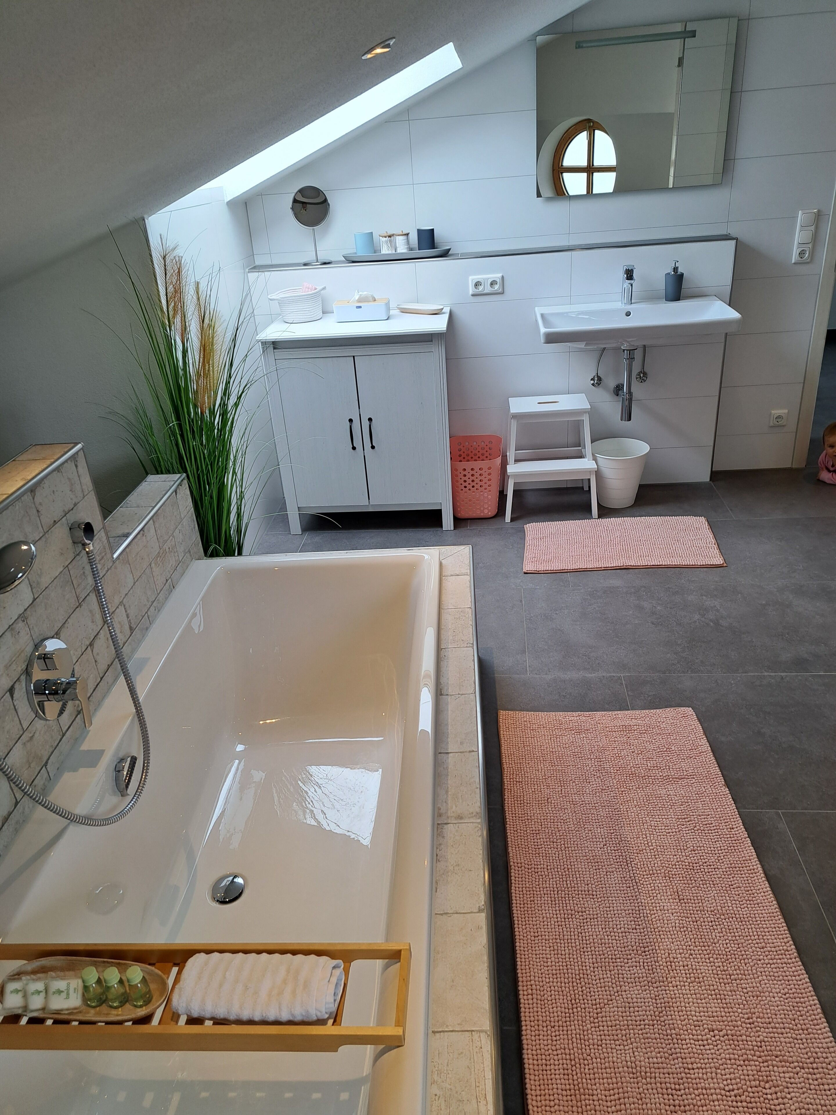 A modern bathroom with a large bathtub, a vanity with a mirror, and a sink. There are towels and toiletries on the counter, and a small step stool is visible.