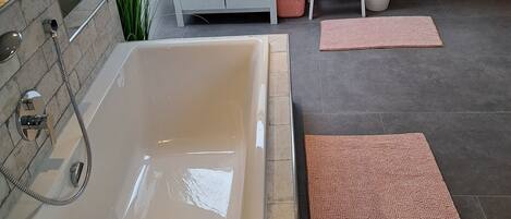 A modern bathroom with a large bathtub, a vanity with a mirror, and a sink. There are towels and toiletries on the counter, and a small step stool is visible.