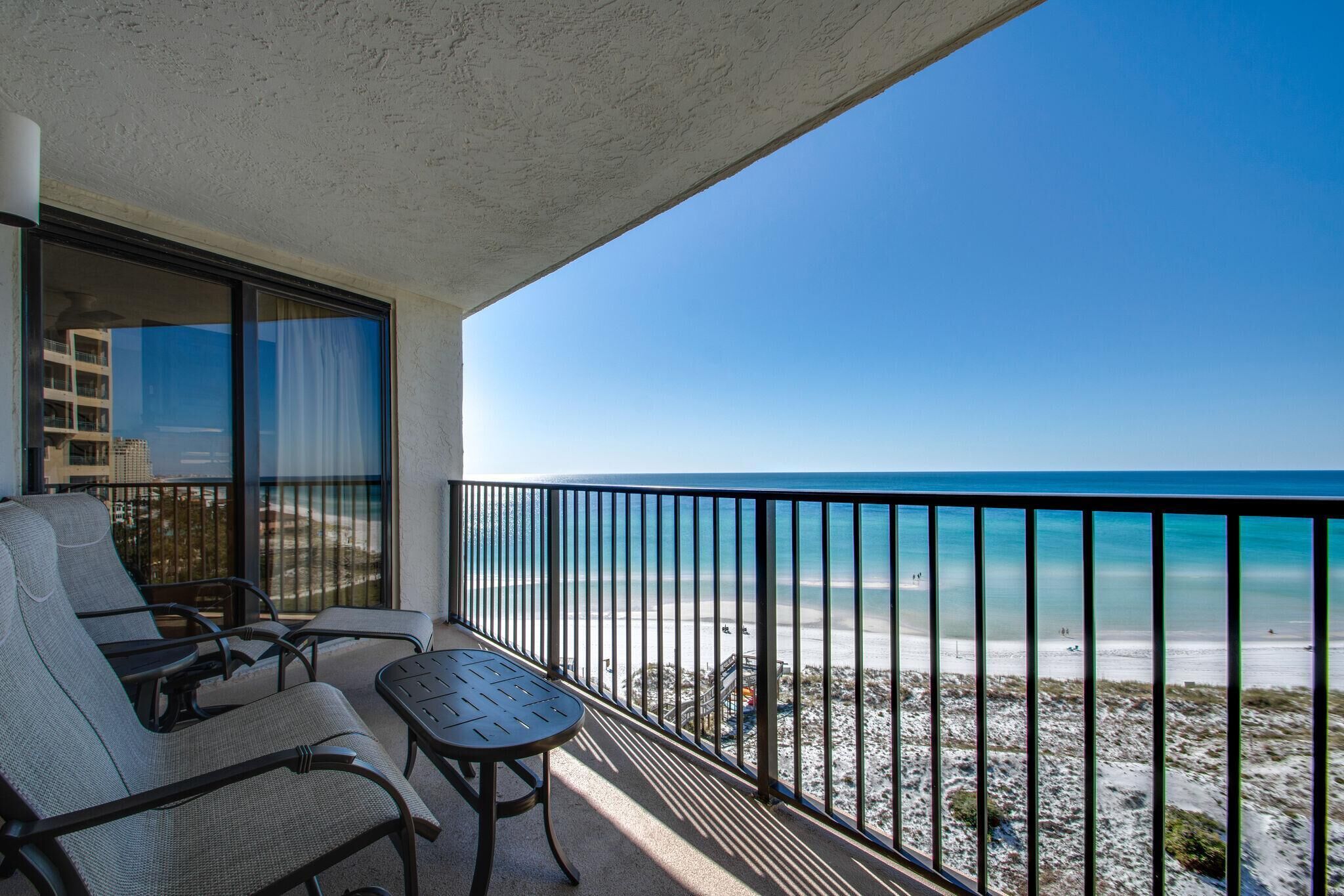 A balcony with a view of the beach, a table, and two chairs.
