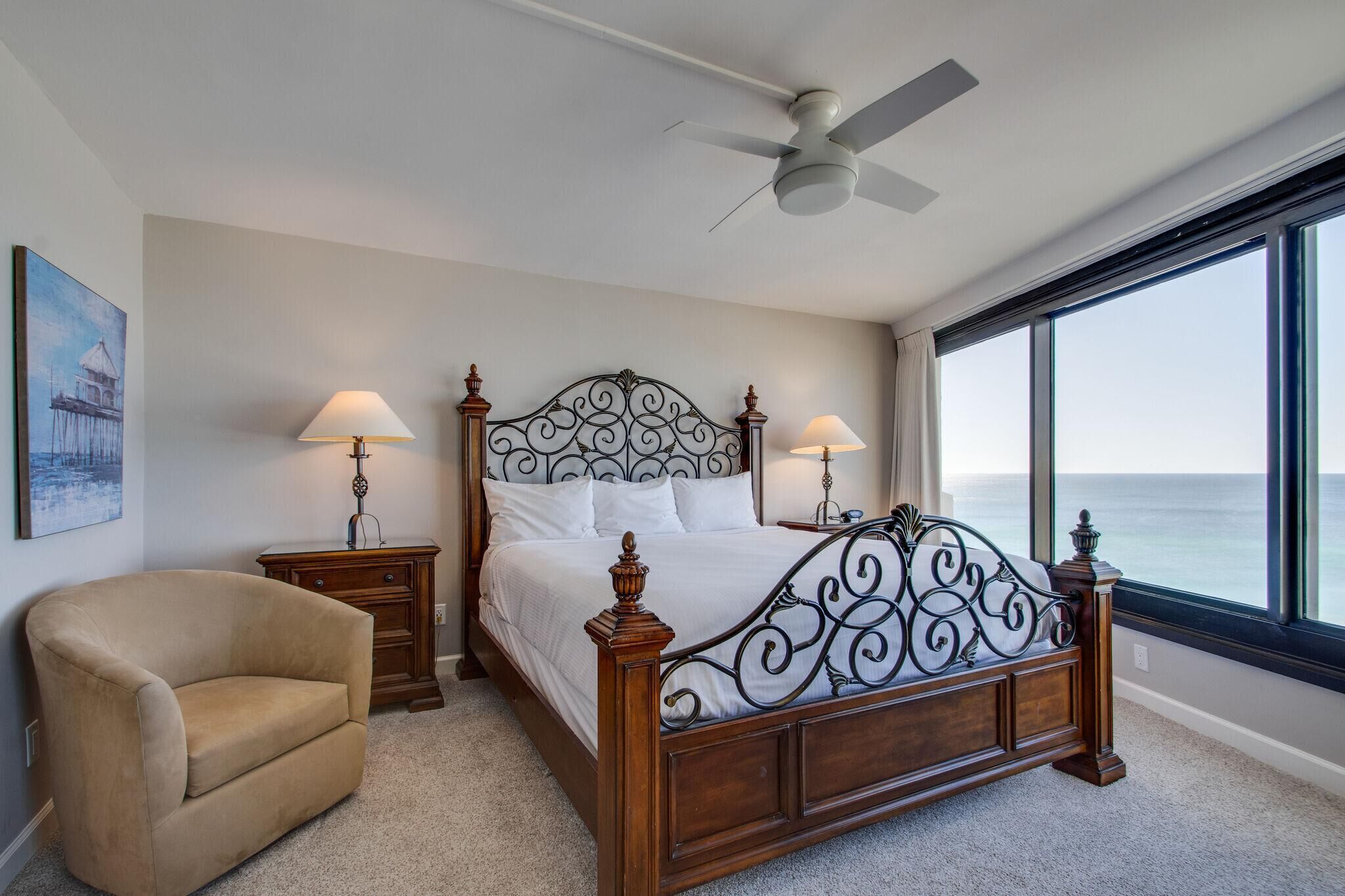 Een slaapkamer met een groot bed, twee nachtkastjes, een stoel, een plafondventilator en een groot raam met uitzicht op de oceaan.