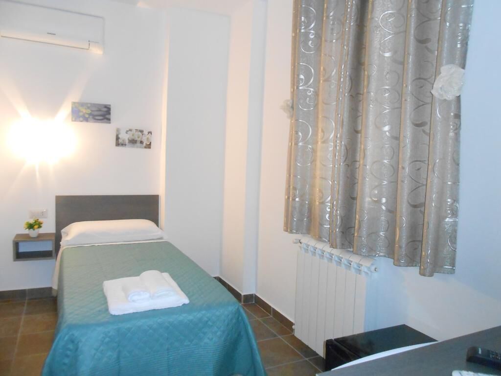 A single bed with a green bedspread and a folded white towel, a bedside table with a small plant, a radiator, and a window with decorative curtains.