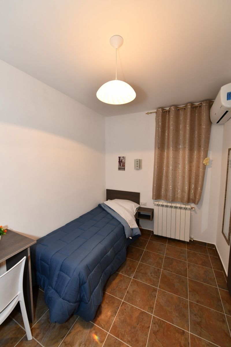 Una stanza piccola con un letto singolo, una scrivania, un termosifone e una finestra con tende.